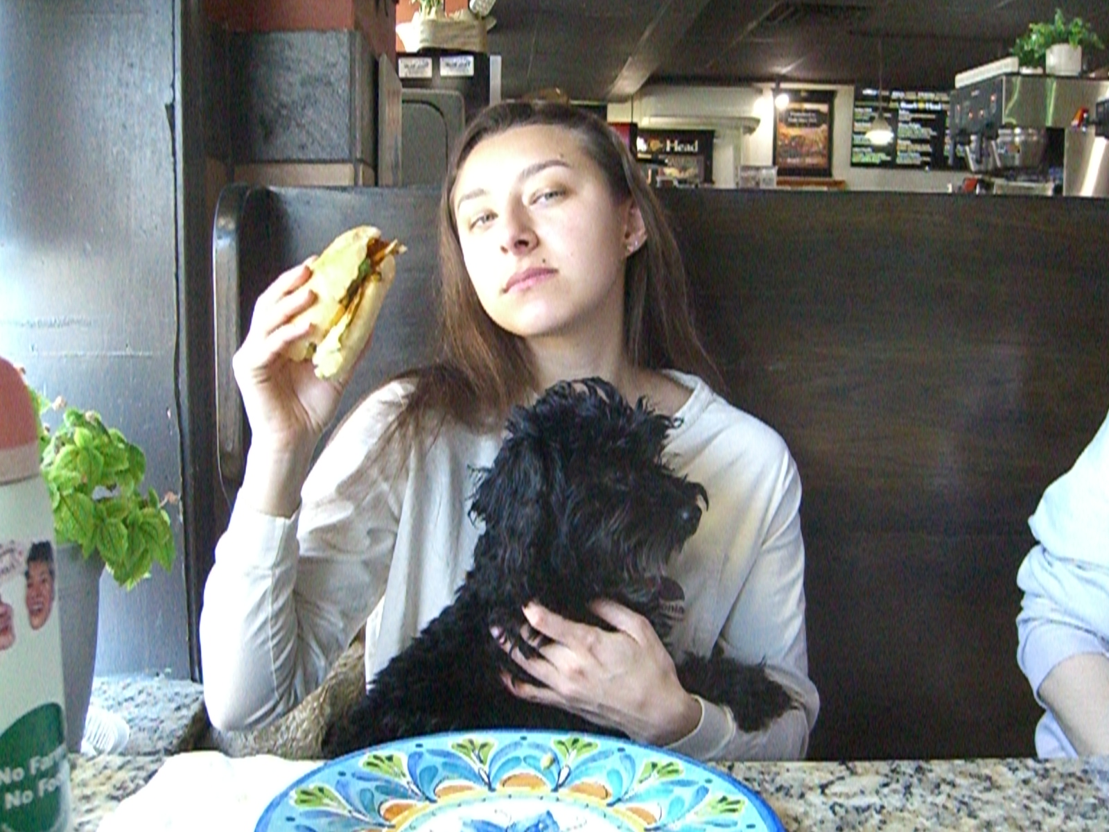 Woman holding a sandwich and a small curly-haired dog at a restaurant