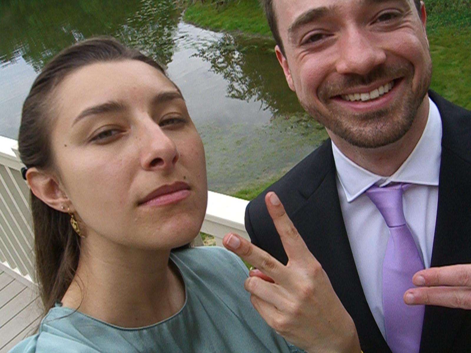 Two people in formal attire throwing peace signs in front of a pond